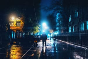 Silhouette of alone stranger in hood at night city street in rain. Creepy killer or stalker, criminal stands in shadow with urban lights reflected in puddles. Thriller horror mysterious atmosphere. This represents stalking.